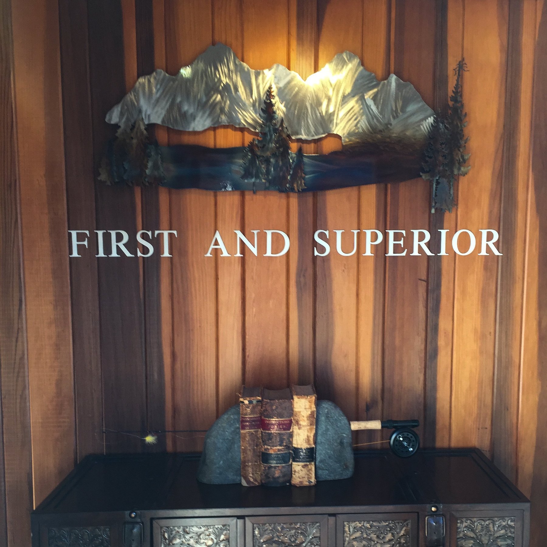 Hand-set letters on the cedar wall of the office reading FIRST AND SUPERIOR, beneath a hammered-brass sculpture of the Selkirk mountains and the lake.